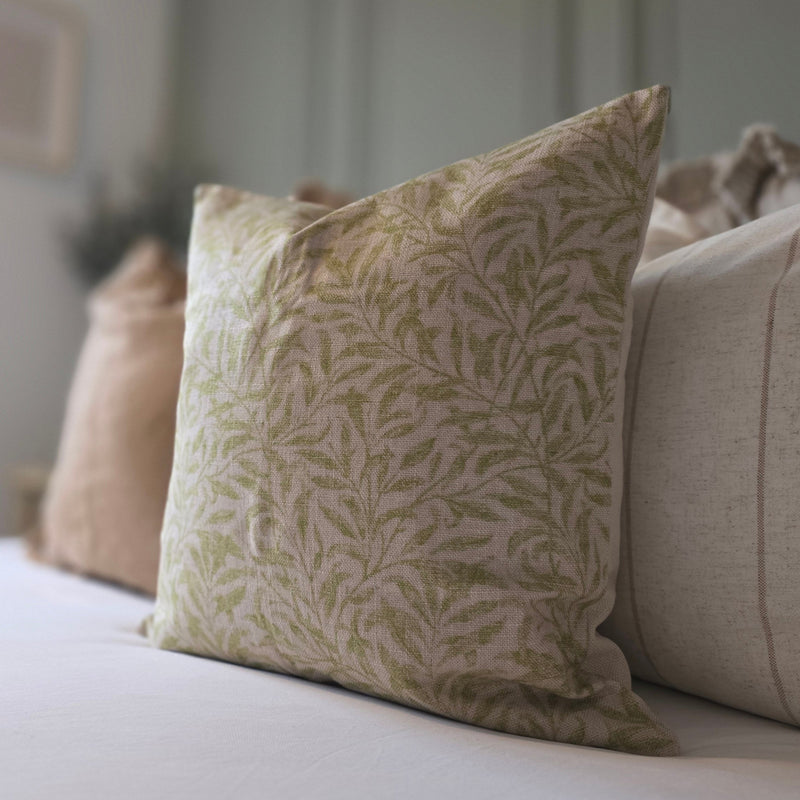 Close up of a square beige cushion with a n all over olive green small leaf print. Sitting on white bedding in front of a set of cream and green cushions