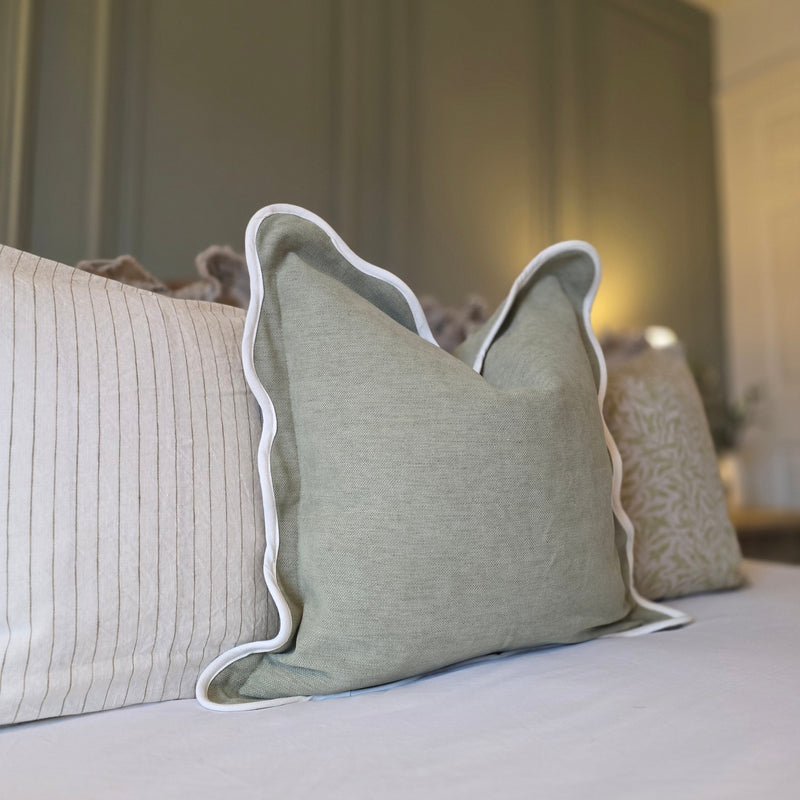 A sage green square cushion with a cream scalloped edge. Sitting on white bedding, with a olive green back wall