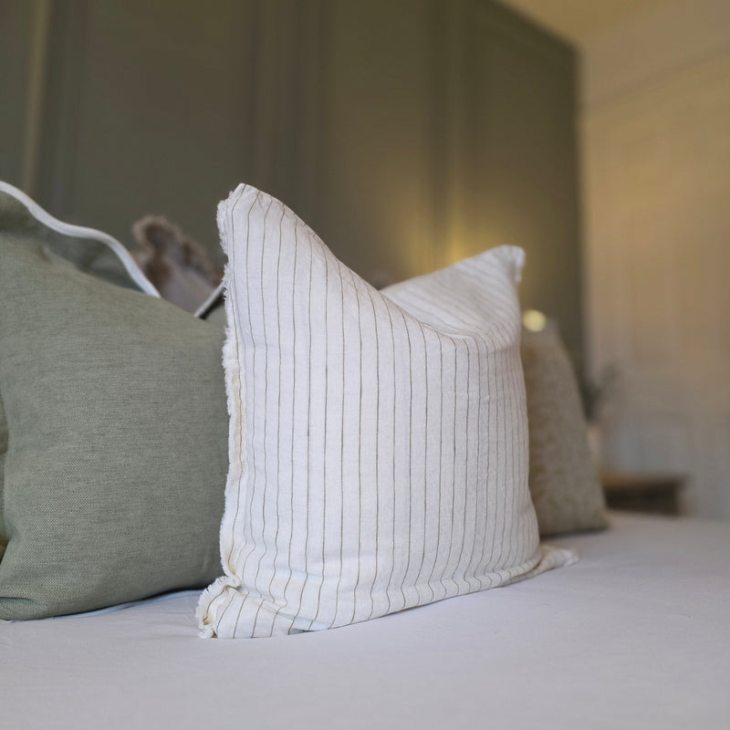 A close up of a square cream cushion with a thin pin stripe olive green vertical all over stripe. Sat on a bed with white bedding and sage green cushions in the background