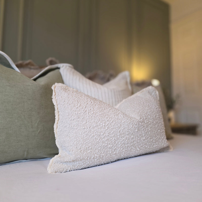 A close up of the cream boucle rectangle cushion laid in front of the set of green cushions on a bed with white bedding and a sage green back wall