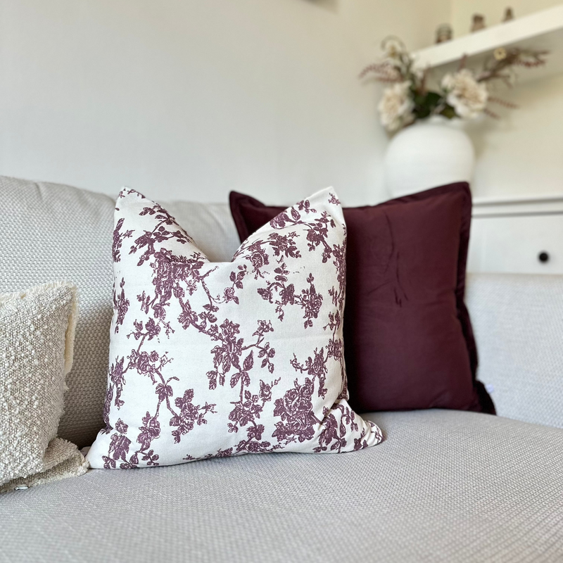 close up of square velvet burgundy cushion and 1 cream square cushion with a burgundy floral print, sat on a cream sofa