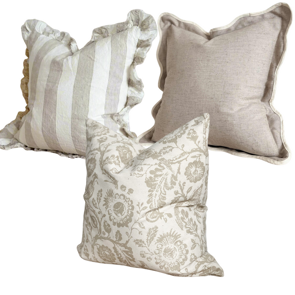 1 square linen ruffled edge cream cushion with a chunky beige vertical repeating stripe. I square beige cushion with a cream piped scalloped edge. 1 square cream cotton cushion with a large brown flower and bird print all over. On a plain white background for contrast.