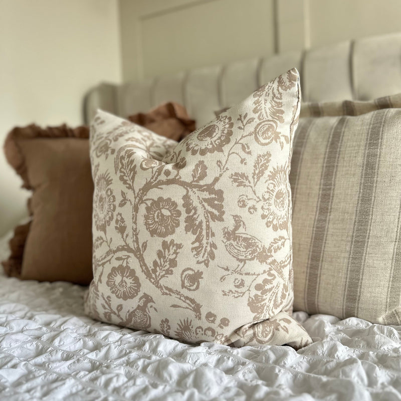 square cream cushion with a large beige floral and bird print. Sat on a bed with white bedding and a beige headboard.