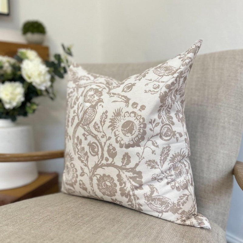 SQUARE CREAM CUSHION WITH A BROWN FLORAL COUNTRY PRINT OF LARGE BEIGE COLOURED FLOWERS, FOLIAGE AND PEACOCK SAT ON A CREAM ARMCHAIR IN A LIVING ROOM