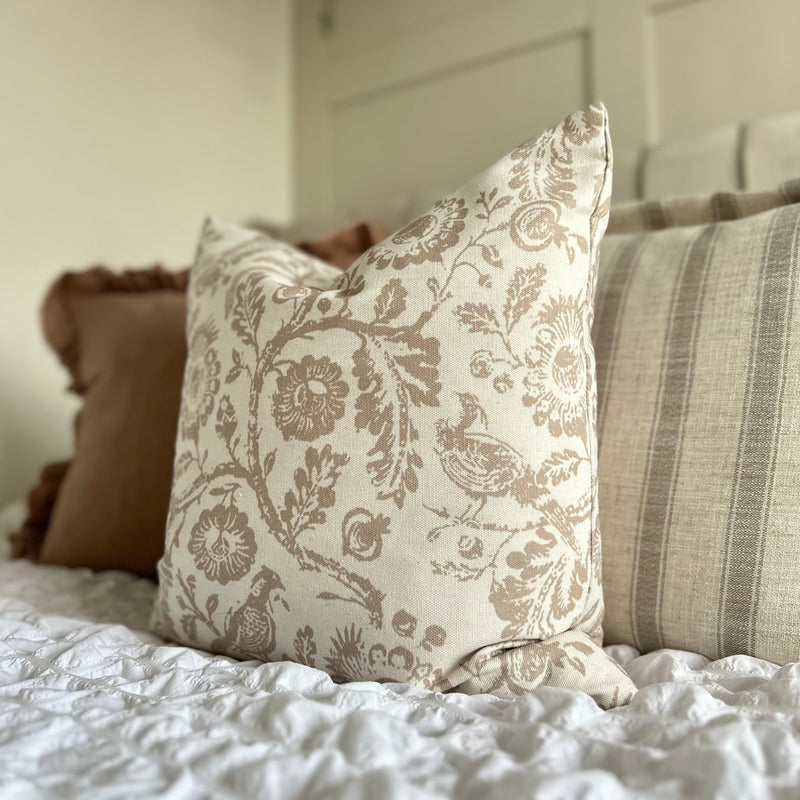 square cream cushion with a large beige floral and bird print. Sat on a bed in a bedroom with white bedding