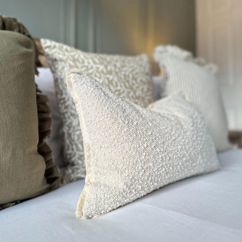 A beige rectangle bouclé cushion cover with a plush texture displayed among other cushions on a bed.