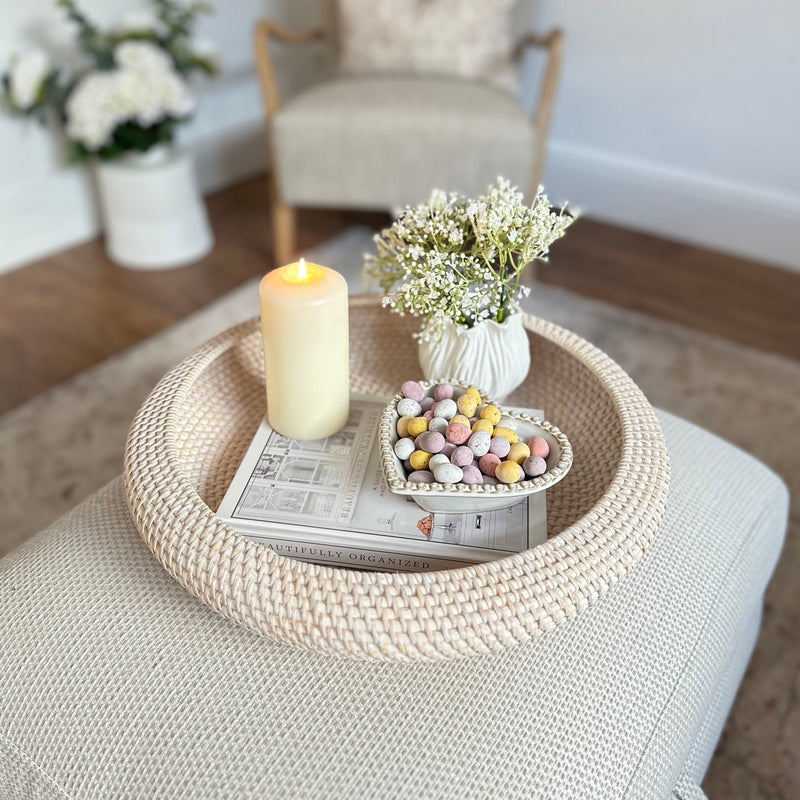 White wash Round Rattan Tray with candle, heart bowl filled with mini eggs and a white vase inside. All sat on a cream footstool