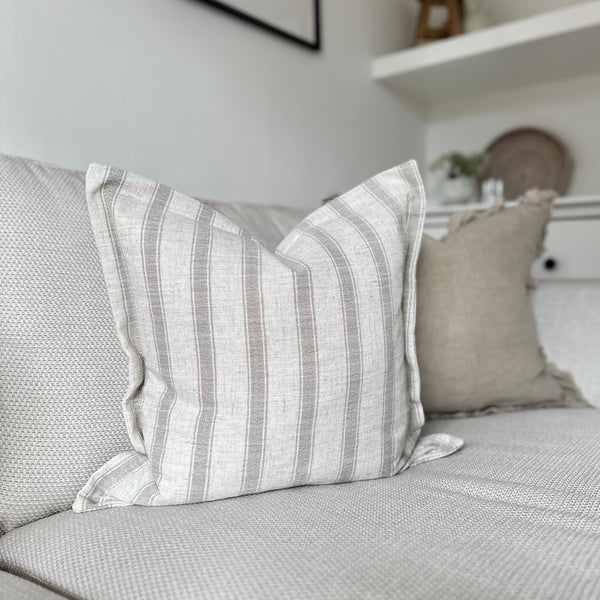 square cream cushion with a thick vertical taupe coloured stripe. Flanged edges. Sat on a cream sofa.