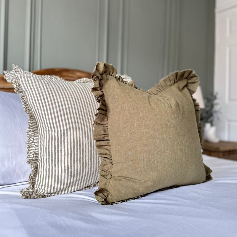 Olive coloured square cushion sat in front of a cream and olive stripe cushion on a bed with white bedding