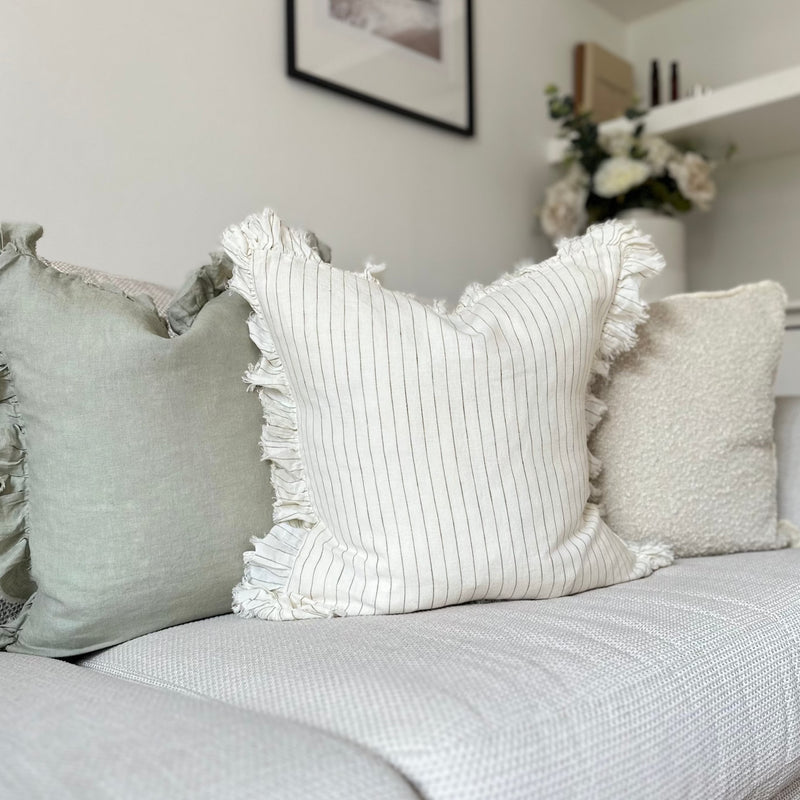 ruffled edge cream square cushion with an olive vertical pin stripe. Sat on a cream sofa in front of a sage green and cream boucle cushion.