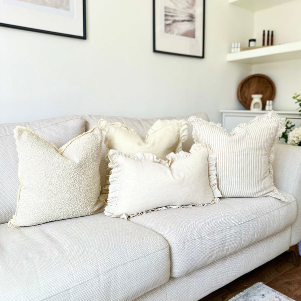 combination of 4 cushions sat on a cream sofa, 3 back cushions one cream ruffled edge with beige vertical repeating stripes. One all over cream with ruffled stripes, one cream boucle with a frayed edge and one rectangle cream cushions with a ruffled edge sat in front