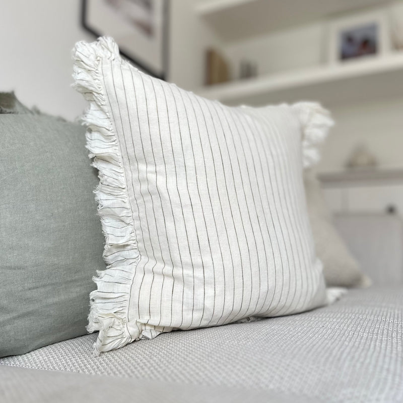 Side profile of a ruffled edge cream square cushion with an olive vertical pin stripe. Sat on a cream sofa.