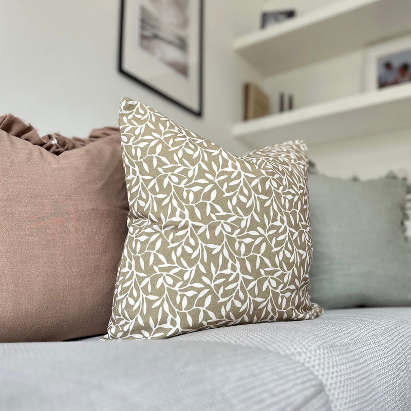 Square cotton olive green cushion with an all over cream coloured olive leaf print. Sat on a cream sofa in front of a brown and green cushion.