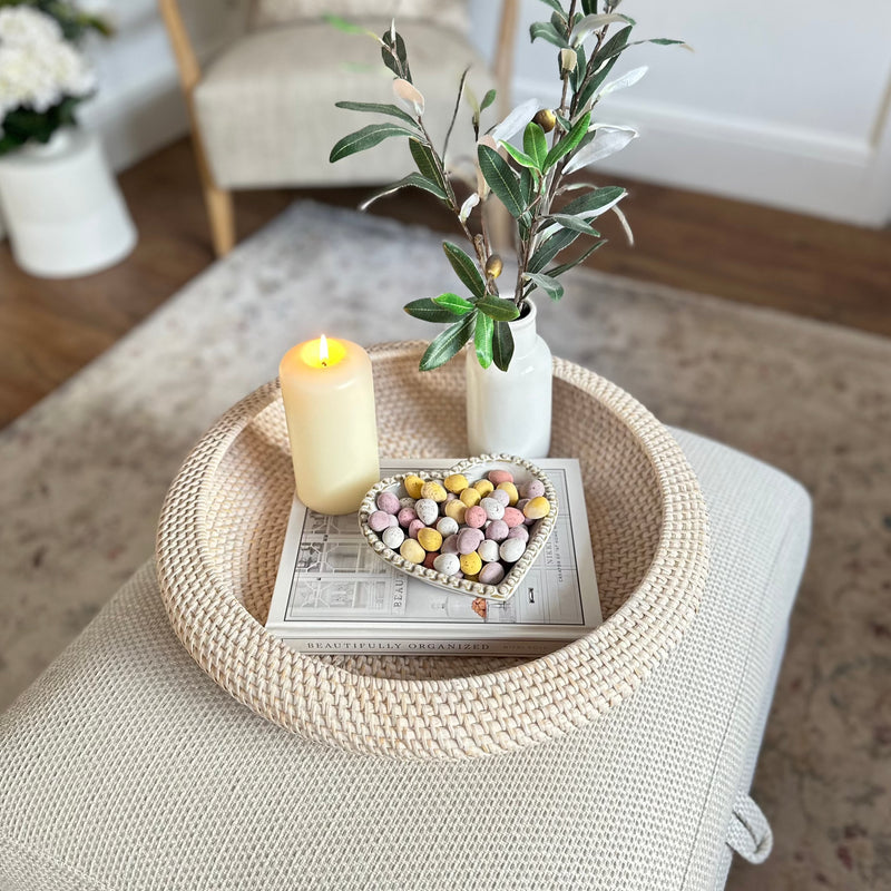 White wash Round Rattan Tray with candle, heart bowl filled with mini eggs and a white vase inside. All sat on a cream footstool