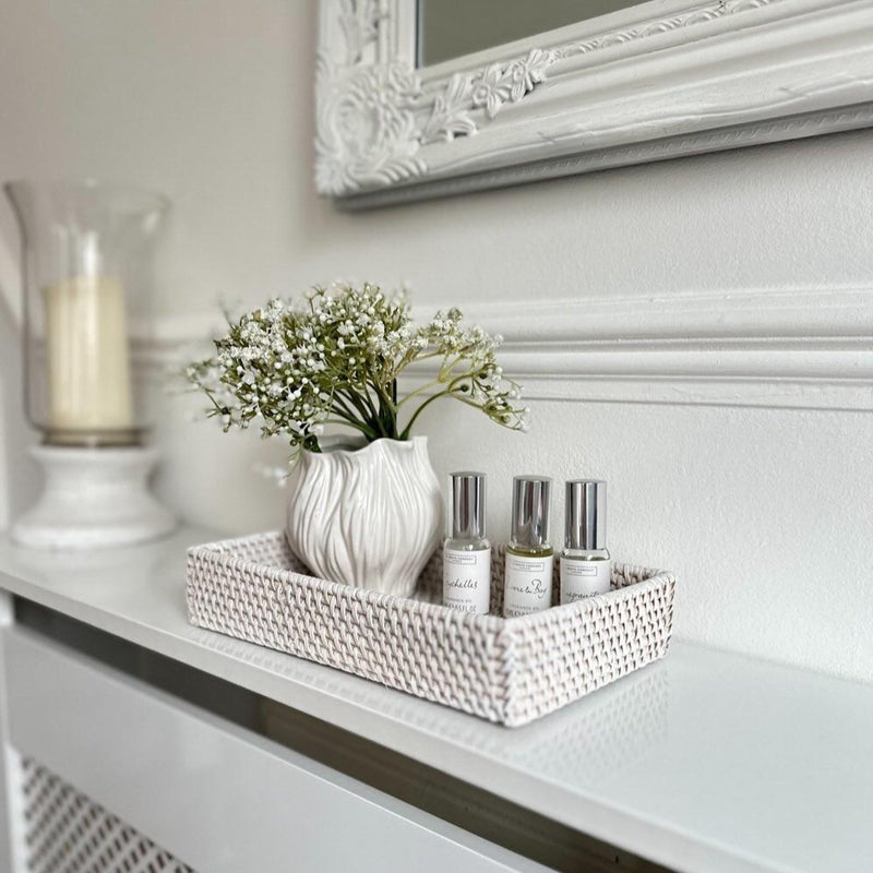small rectangle whitewash rattan tray. Displayed with a white floral shaped vase and small oil fragrance bottles. sat on a white radiator cover