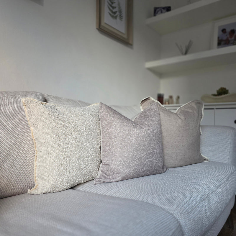 Set of 3 cushions. one square greige with a cream piped scalloped edge, one square greige cushion with an all over greige leaf print, 1 square cream boucle cushion all sat on a cream sofa.