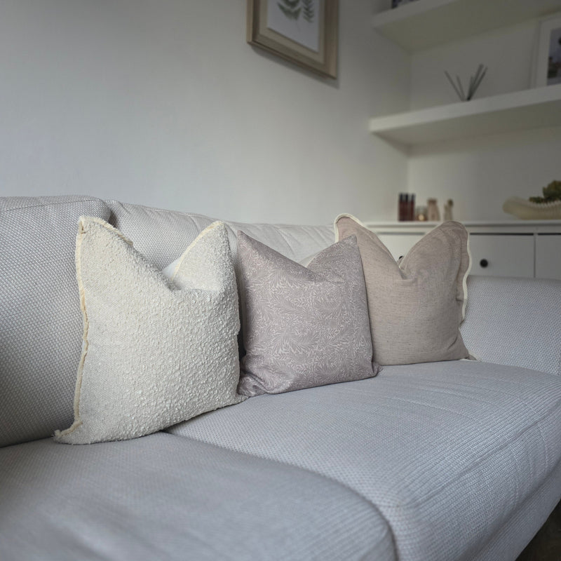 Set of 3 cushions. one square greige with a cream piped scalloped edge, one square greige cushion with an all over greige leaf print, 1 square cream boucle cushion all sat on a cream sofa