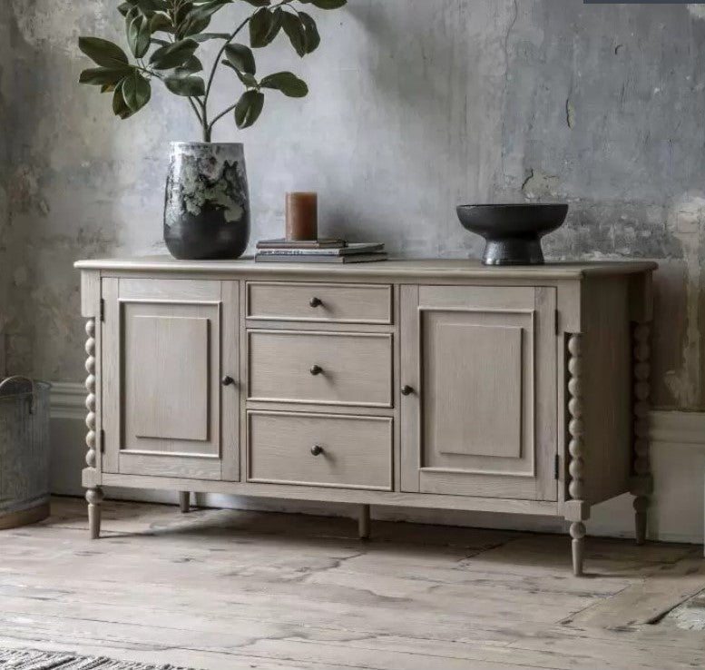 Oak Bobbin 2 Door 3 Drawer Wooden Sideboard sat in a moody living room setting. Displaying vases, books and candles on top for display