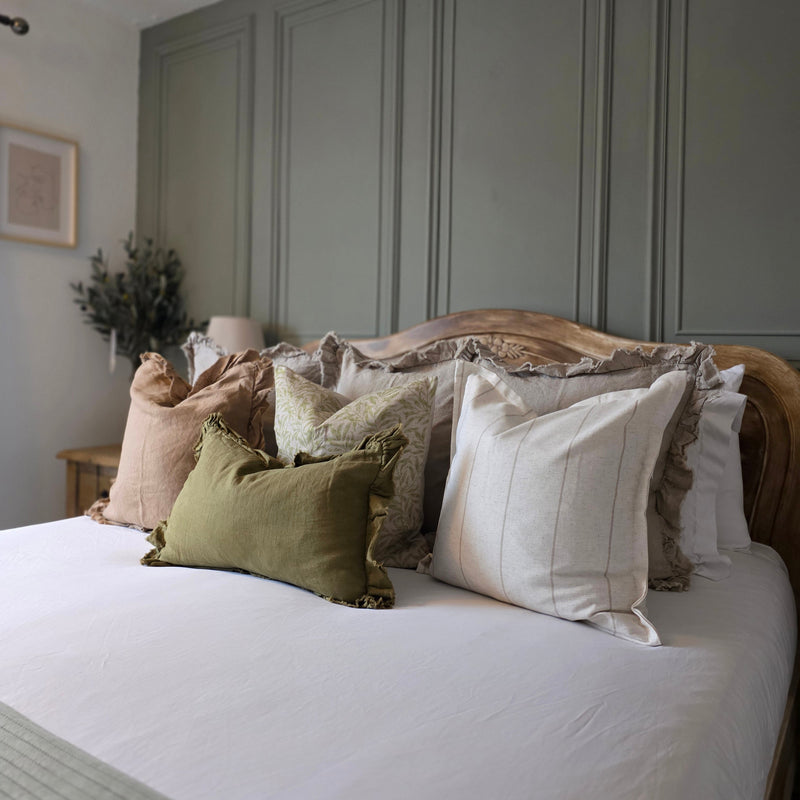 Set of 4 cushions. 1 square brown linen cushion with ruffled edge. 1 square beige cushion with an all over green leaf print. 1 square cream cushion with a repeating slim beige vertical stripe with a flanged edge. 1 rectangle olive green linen cushion with a ruffled edge. All sat on a bed with white bedding and a panelled olive green back wall.