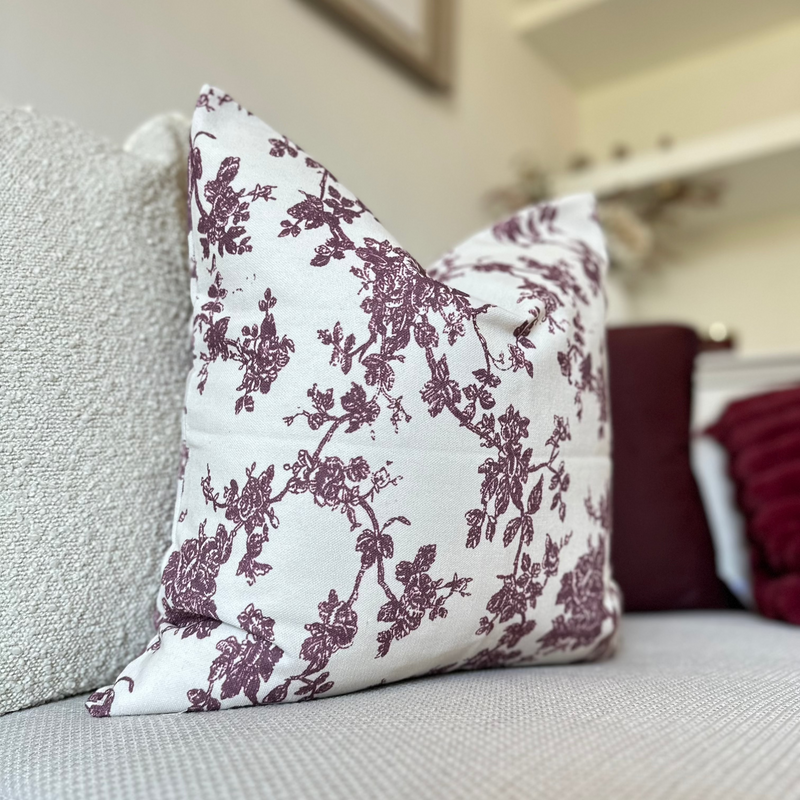 Square cream cushion with an all over plum floral print sat in front of a velvet plum cushion on a cream sofa