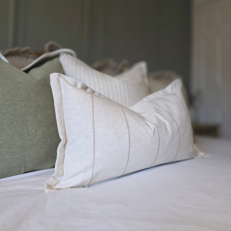 A rectangle cushion with a flanged edge. Cream, stone colour with a subtle vertical beige stripe repeating from left to right. Sat on a bed with white bedding