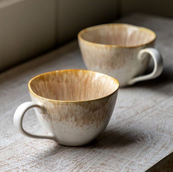 Caramel colour cute set of 2 mugs with an imperfect round shape and a cute handle.  2 tone pink and cream more brown on top blending to a soft cream on the bottom. Both sat on a wooden worktop