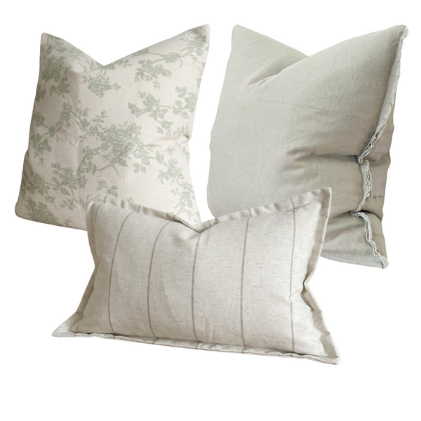 Set of 3 cushions. 1 square cream cotton cushion with sage green floral print. 1 square weathered edge sage green cushion. 1 cotton rectangle stone cream cushion with a vertical beige stripe and flanged edge. On a white plain background.