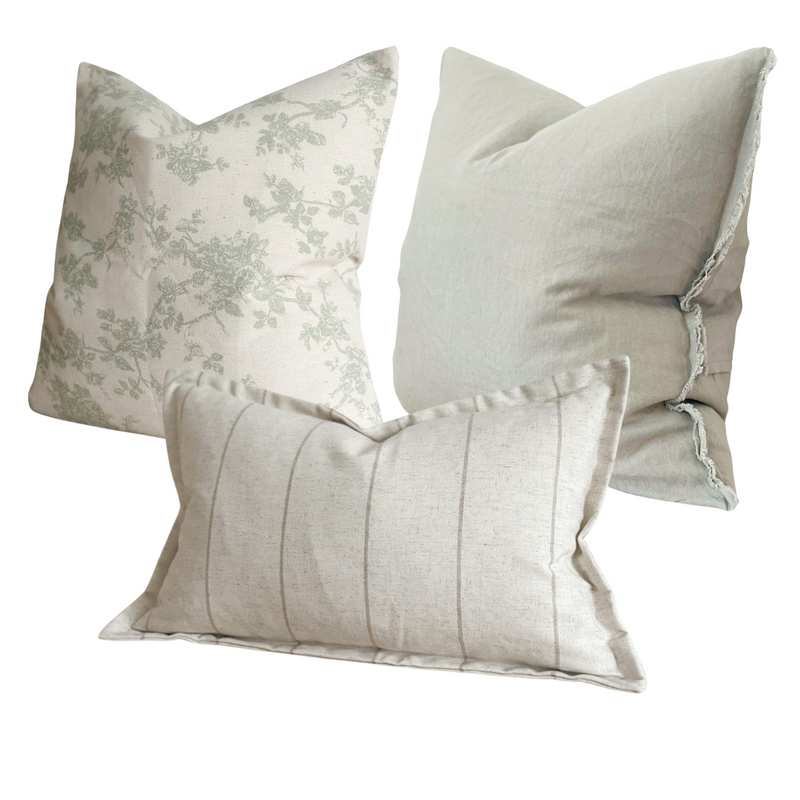 Set of 3 cushions. 1 square cream cotton cushion with sage green floral print. 1 square weathered edge sage green cushion. 1 cotton rectangle stone cream cushion with a vertical beige stripe and flanged edge. On a white plain background.