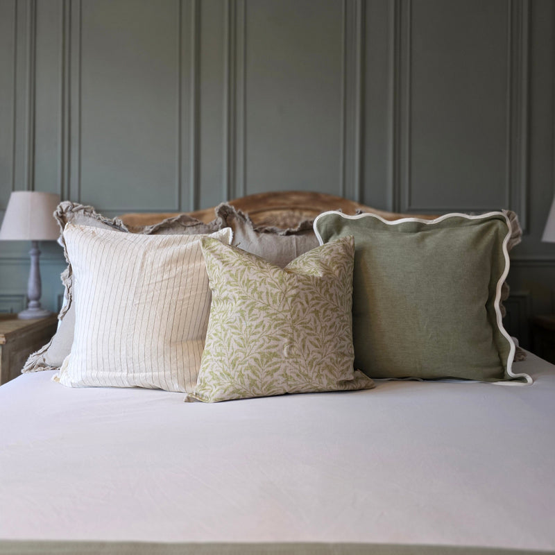 A set of 3 cushions. 1 sage green with a cream scalloped edge, 1 beige with an all over intricate green leave print and 1 cream with an olive vertical all over pin stripe. Sat on a bed with white bedding and an olive green back wall
