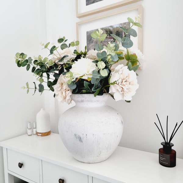 white vase filled with faux white and cream hydrangeas and eucalyptus. Sat on a white sideboard in a living room