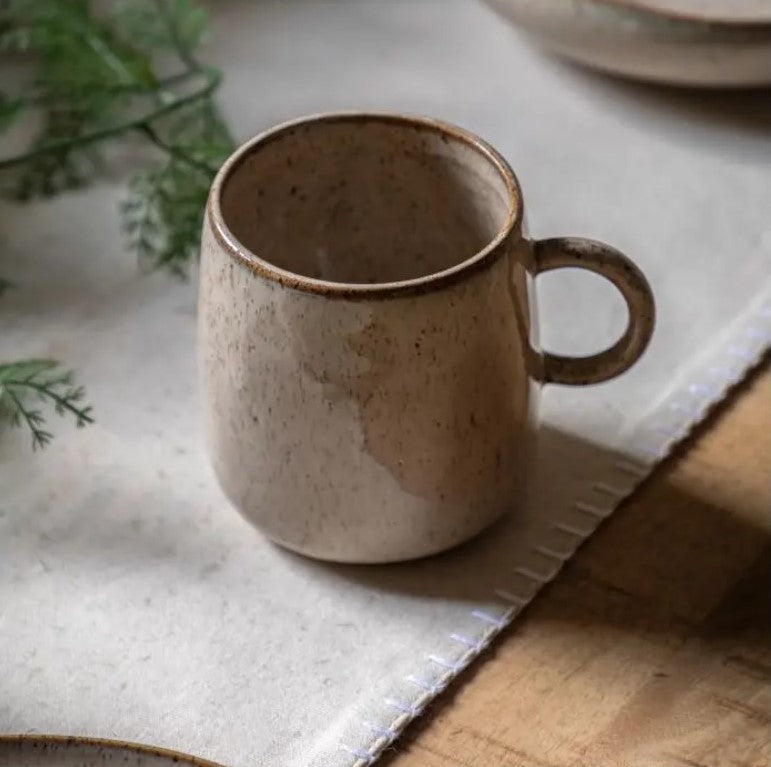 Speckled Glazed Mug Oatmeal coloured with a darker brown rim sitting on a beige linen table cloth in the centre of a wooden dining table