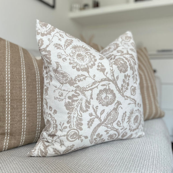 square cream cotton cushion with a large brown floral all over pattern with flowers, leaves and peacocks in a brown/beige. Sat on a cream sofa.