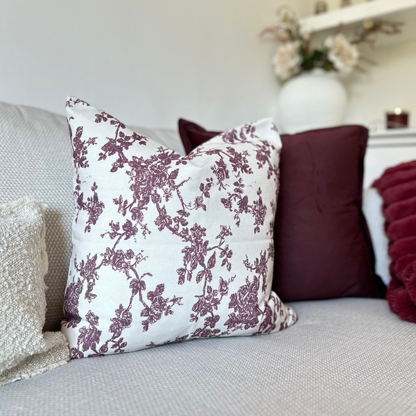 Square cream cushion with an all over plum floral print sat in front of a velvet plum cushion on a cream sofa