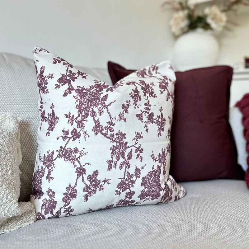 Square cream cushion with an all over plum floral print sat in front of a velvet plum cushion on a cream sofa