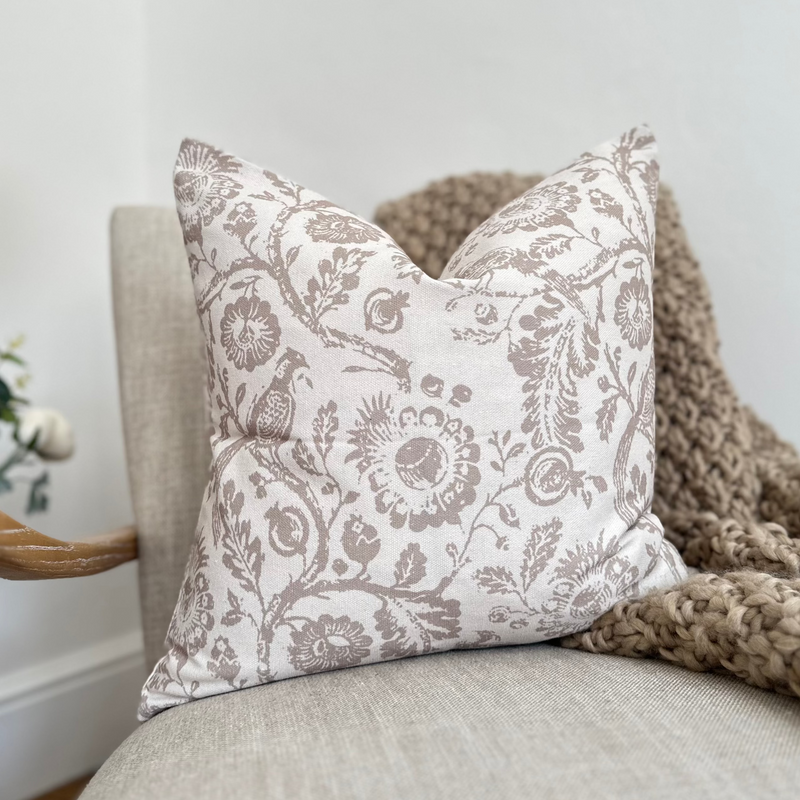 Square cream cotton cushion with a large brown floral and bird print front and back. Cushion sat on a cream armchair with a brown chunky knit thrown behind.