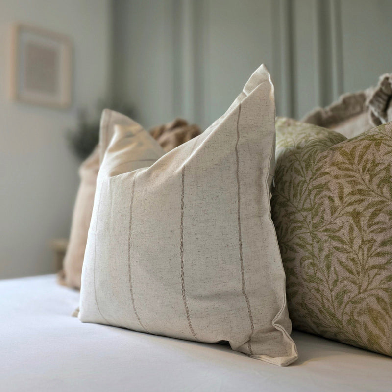 Square flanged edge cotton cream and beige stripe cushion sat on a bed with white bedding