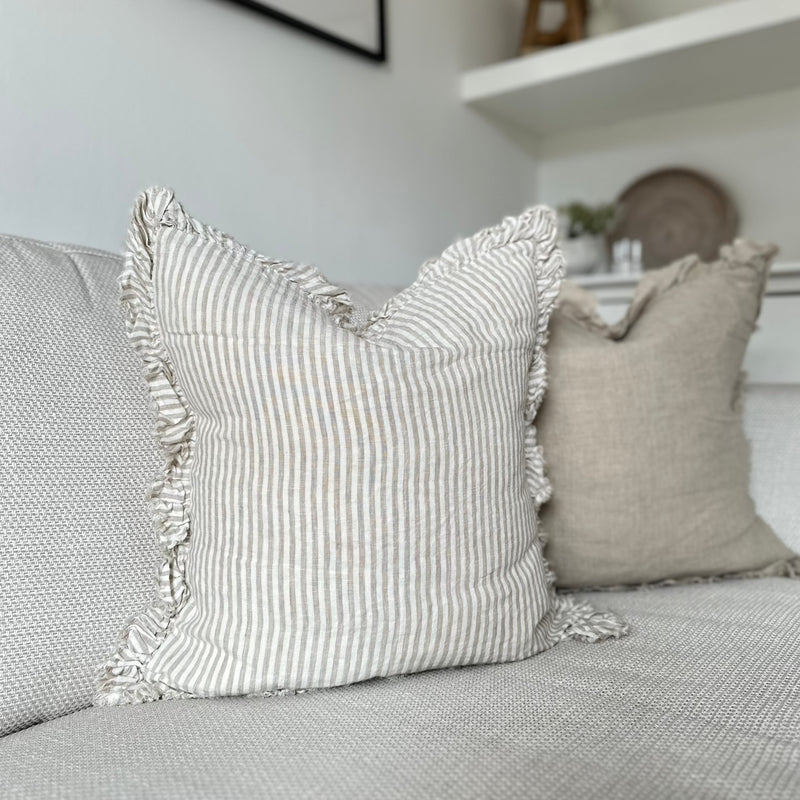 Square ruffled edge cream linen cushion with an vertical close repeating beige stripe. Sat on a cream sofa.