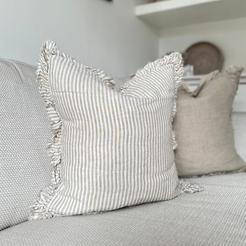 Square ruffled edge linen cream cushion with an vertical close repeating beige stripe. Sat on a cream sofa.