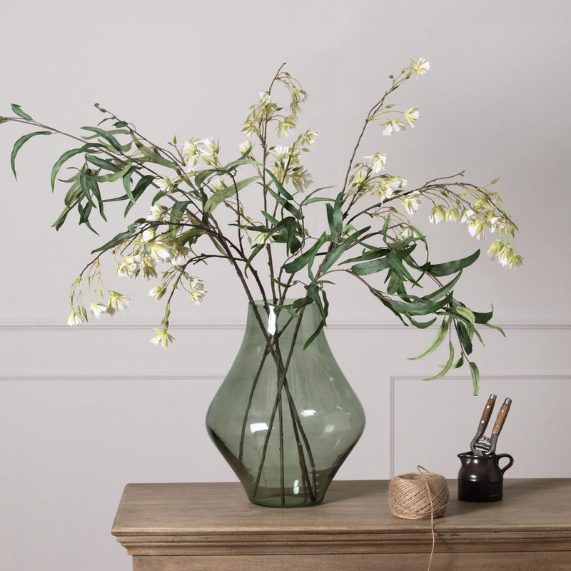 Stamford Sage Green Clear Glass Vase styled with wispy green stems and white flowers sat on a wooden sideboard against a white wall