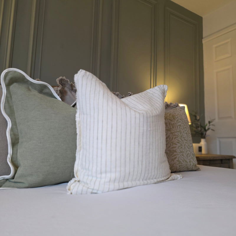 Close up of set of 3 Sage green and cream cushions. Weathered edge cream linen cushion with a vertical, all over olive pinstripe, sat on a bed in front of green cushions