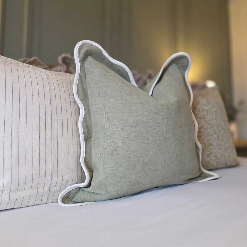 Close up of sage green cushion with a cream scalloped edge. Sat on a white bedding