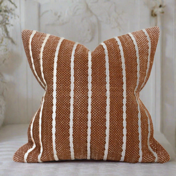 square auburn cushion with vertical repetitive stripes sat in a white bedroom with white sheets.