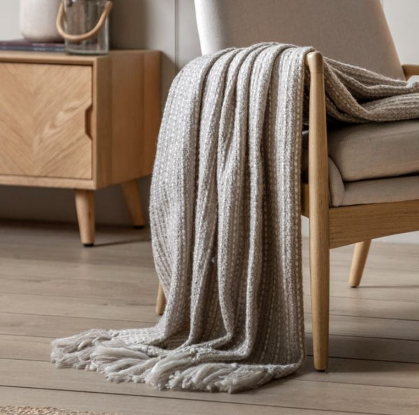 A soft grey throw with a subtle cream stich throughout with chunky tassels along the bottom, thrown over a cream chair for styling