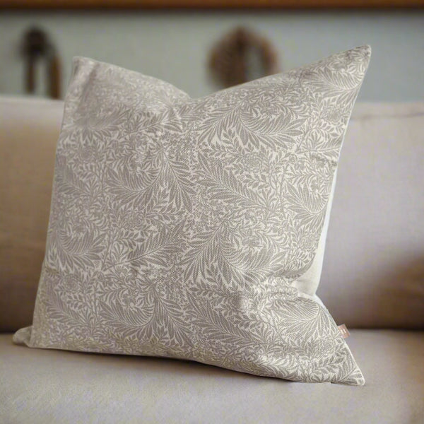 square cushion with an intricate greige floral print on a cram background. Sitting on a cream sofa