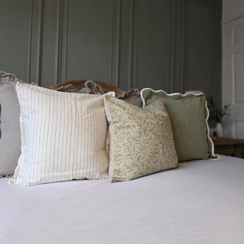 Side angle of a set of 3 cushions. 1 sage green with a cream scalloped edge, 1 beige with an all over intricate green leave print and 1 cream with an olive vertical all over pin stripe. All sat in a bedroom setting with white and green accents