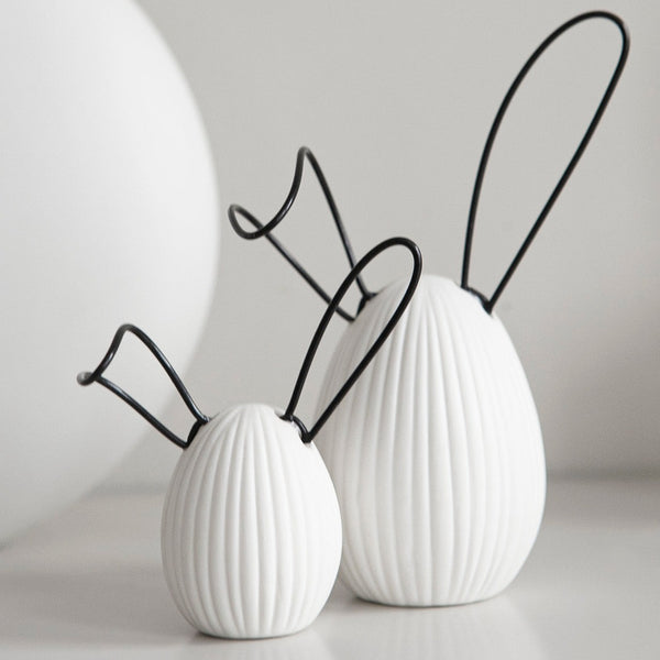 A decorative white egg with a ribbed design and black wire ears, adding a playful touch to any space. The unique design and color scheme make it a versatile addition to any decor style, perfect for Easter or springtime. Ideal for display on shelves, mantels, or desks, and makes a great gift for friends, family, or co-workers.