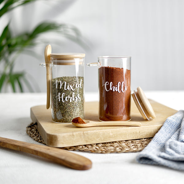 Two round glass spice jars with an eco friendly bamboo lid and serving spoon, with custom personalised wording for Mixed Herbs and Chilli