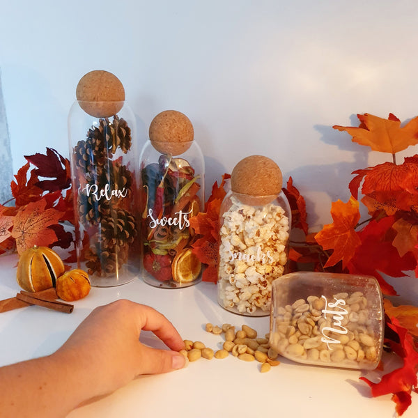 Three glass jars with cork stoppers, each different variants of size, with custom personalised wording on the face of the jar, all containing beautiful autumn supplies.